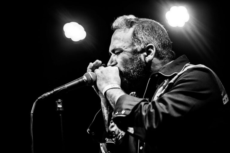Een man speelt mondharmonica op een podium, omgeven door felle lichten.