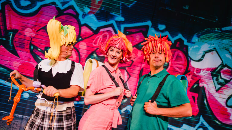 Twee acteurs met grote rode pruiken en rugzakken om. 1 acteur met een gele pruik