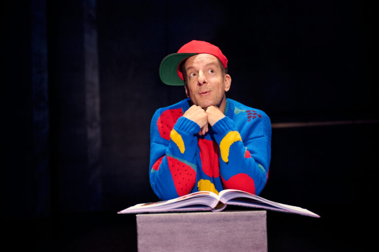 Een vrolijke man in een kleurrijk gebreid vest en een pet, die met een glimlach leunt op een open boek.