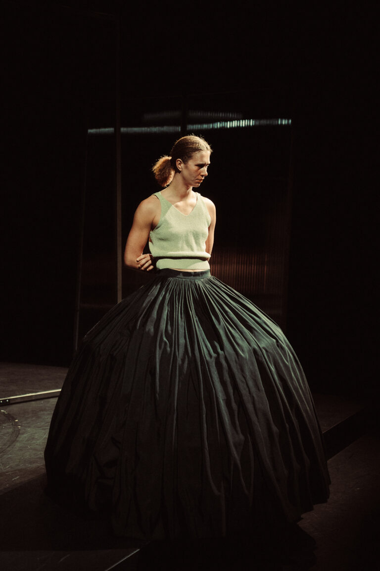 Een vrouw in een stijlvolle, volumineuze zwarte rok en een lichte top staat in een theatrale setting.