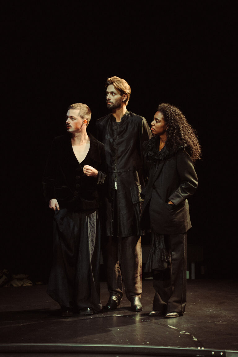 Drie acteurs in donkere kleding staan op een theaterpodium met een zwarte achtergrond.