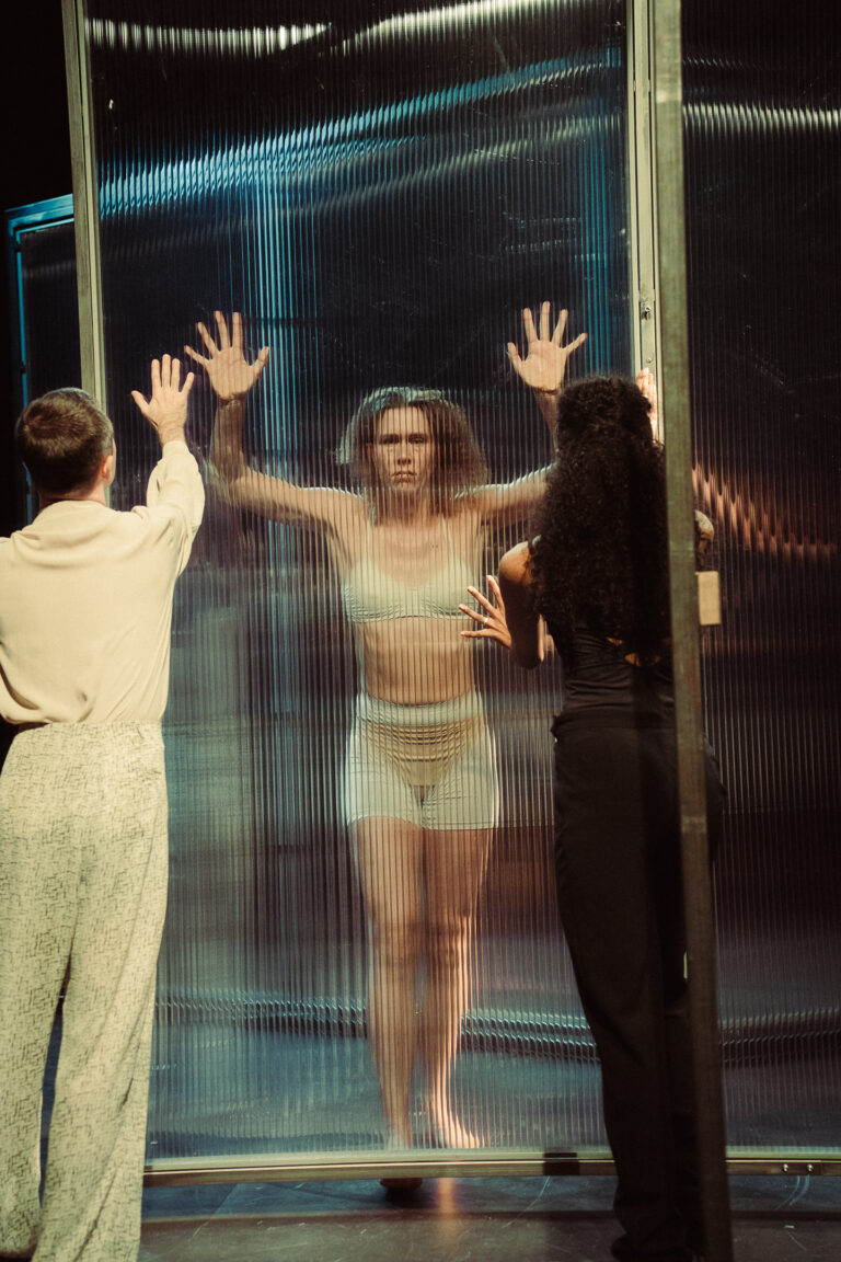 Een vrouw staat achter een glazen wand met opgestoken handen, terwijl twee mensen aan de andere kant staan.