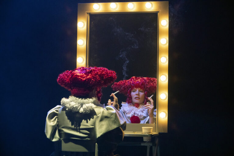 Actrice met rode pruik en nepsigaret voor een spiegel.