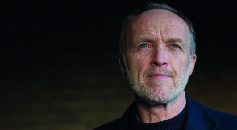 Een contemplatief portret van een middelbare man met een baard, gekleed in donkere kleding, staand tegen een zacht verlichte bakstenen muur.