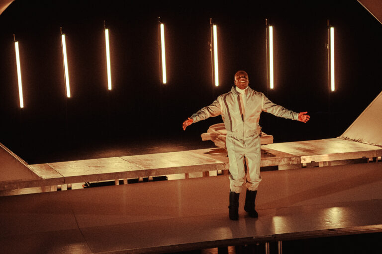 Een performer in een glanzend kostuum staat mid-actie op een podium met fel gemaakte lichtstrips op de achtergrond.