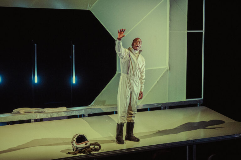 Een astronaut in een witte ruimteoveral staat op een podium en zwaait, met een helm en futuristische achtergrond.