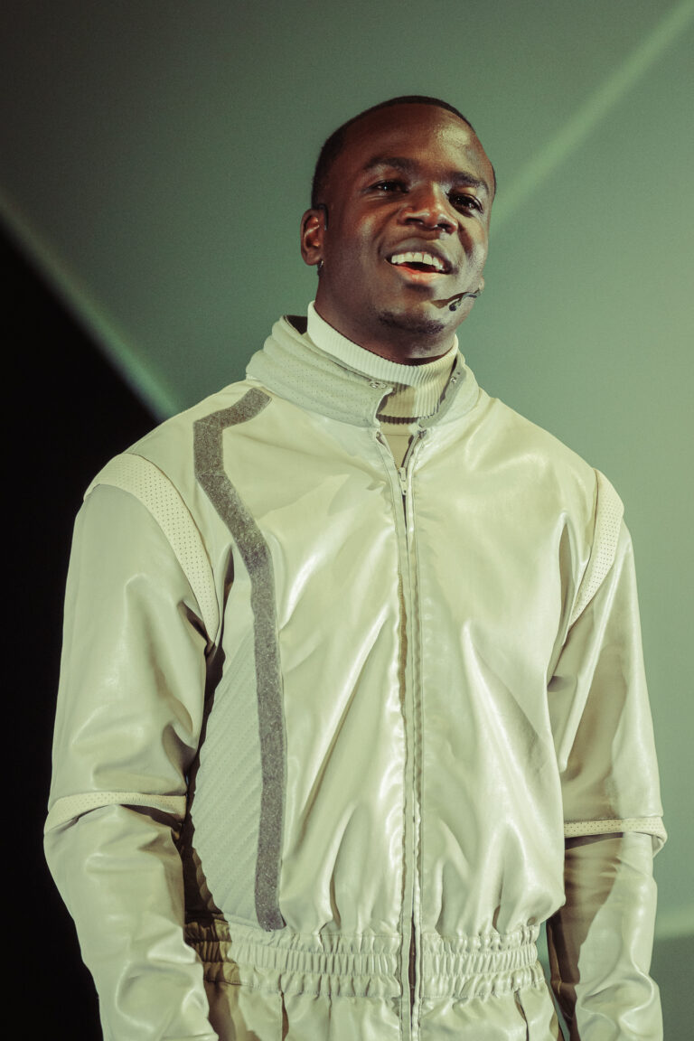 Een persoon met een glimlach draagt een lichte, modieuze jas en staat in een lichte omgeving.