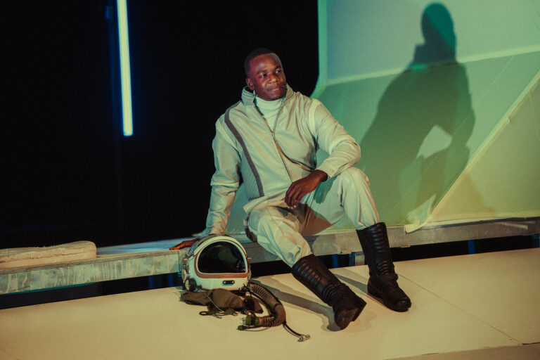 Een persoon in een ruimtepak zit op een platform naast een astronauthelm, omringd door kleurrijke verlichting.