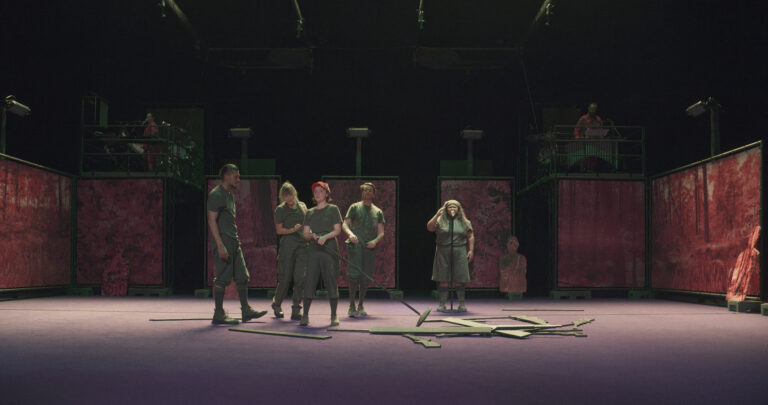 Een groep acteurs in groene outfits staat op een podium omringd door schermen en afgebroken rekwisieten.