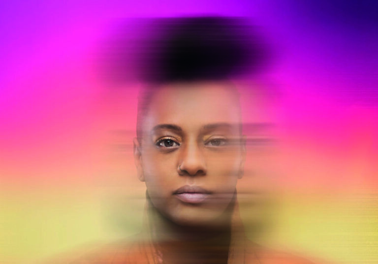 Een persoon met een korte, donkere haarsnit staat voor een kleurrijke, vervaagde achtergrond.