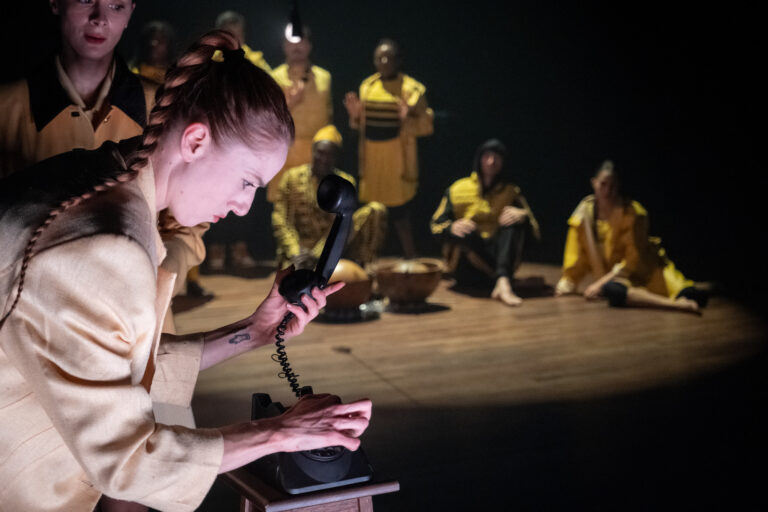 Een vrouw met een telefoon in de hand staat voor een groep mensen die in gele kleding zitten en luisteren.