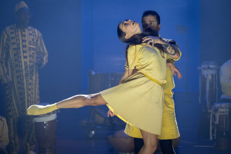 Een dansend paar in gele outfits voert een emotionele choreografie uit op een blauwe achtergrond.