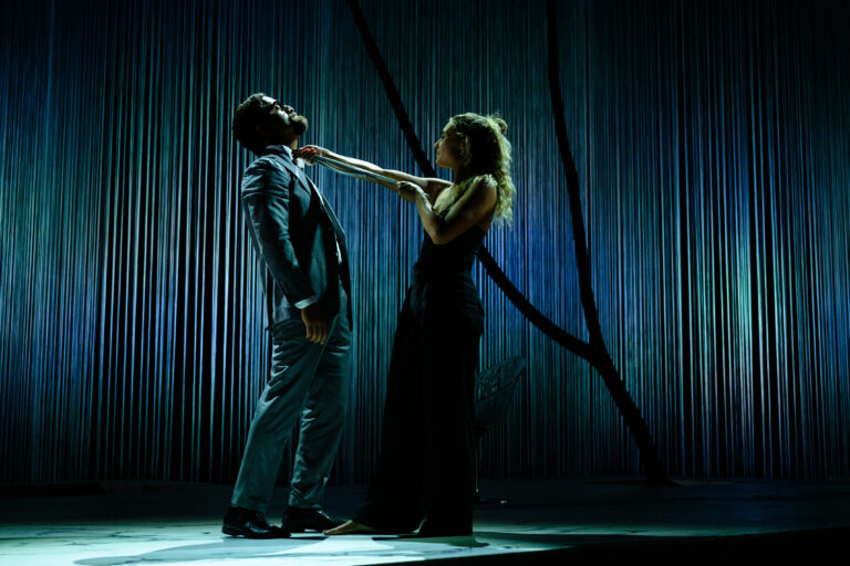 Een vrouw in een zwarte jurk trekt aan de kraag van een man in een grijs pak, met een dramatische achtergrond.