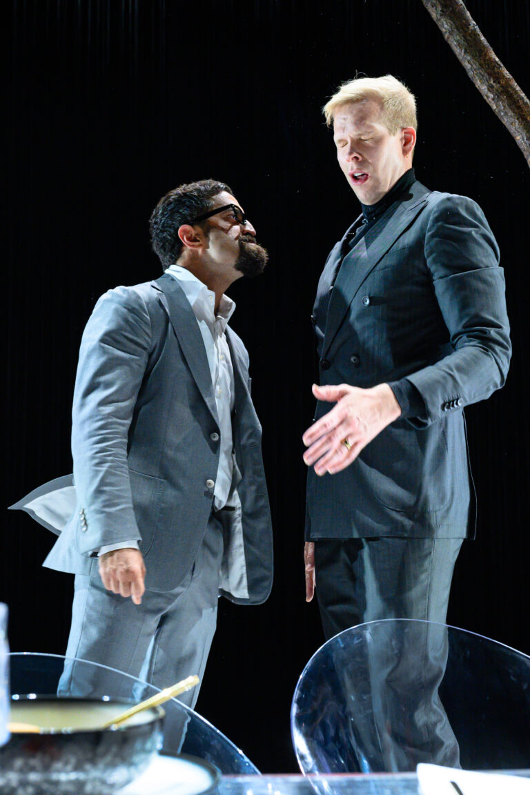 Twee mannen in formele kleding staan tegenover elkaar op een podium, met één man die zijn hand uitsteekt.