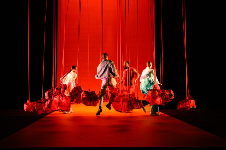 Een groep dansers in kleurrijke kleding performt op een podium met een felrode achtergrond.