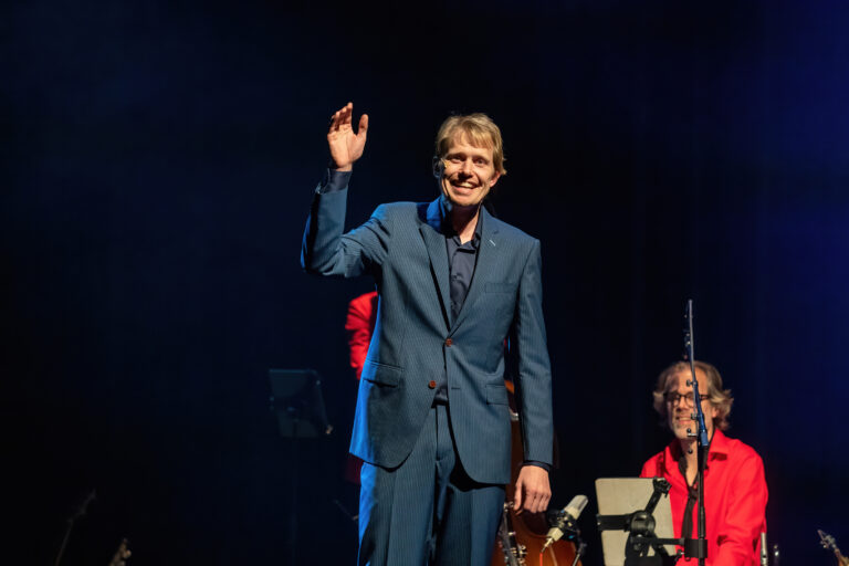 Een man in een blauw pak steekt zijn hand op tijdens een optreden, met een muzikant op de achtergrond.