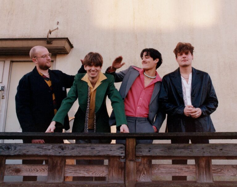 De vier bandleden van The Vices die op een houten balkon staan. Twee kijken in de camera en twee leden kijken naar elkaar.