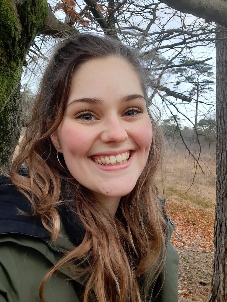 Jonge vrouw met een glimlach, staand in een bosrijke omgeving met kale bomen op de achtergrond.