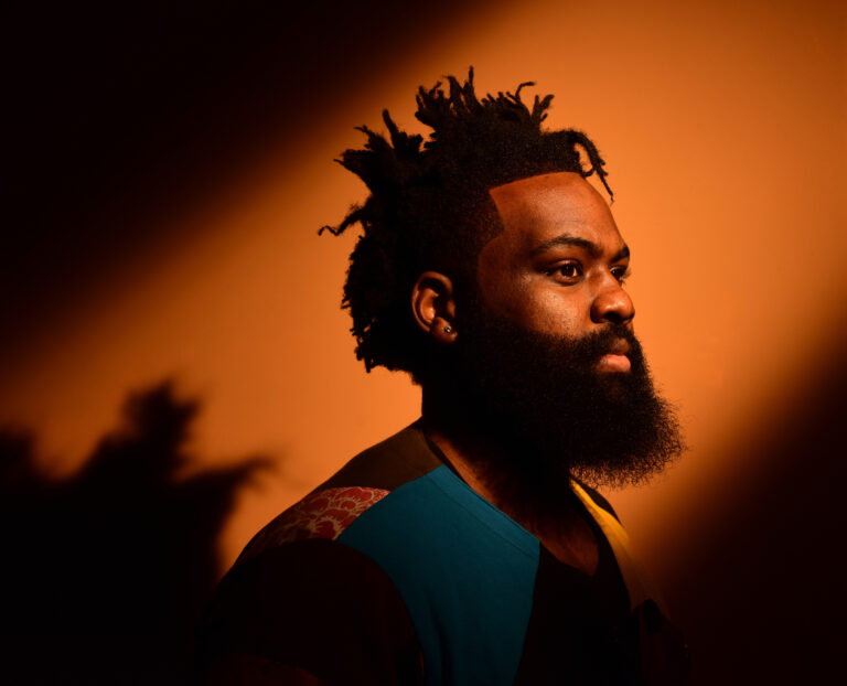 Een man met een volle baard en kroeshaar staat in profiel voor een warme oranje achtergrond.