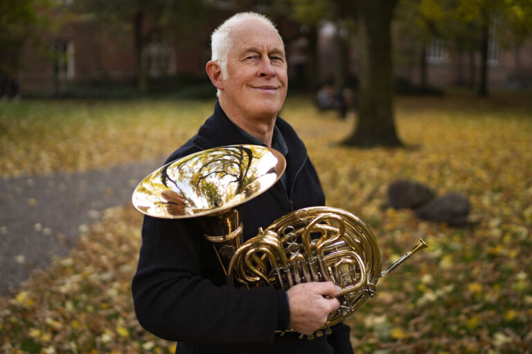 Een man staat buiten in een omgeving vol herfstbladeren en houdt een glanzende hoorn vast.