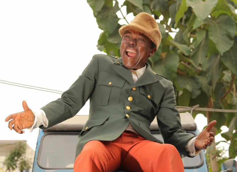 Een vrolijke man in een groen jasje en hoed, zittend op een voertuig met een glimlach en uitgestoken armen.