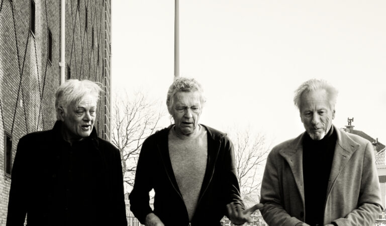 Drie mannen lopen samen buiten in een zwart-wit foto.
