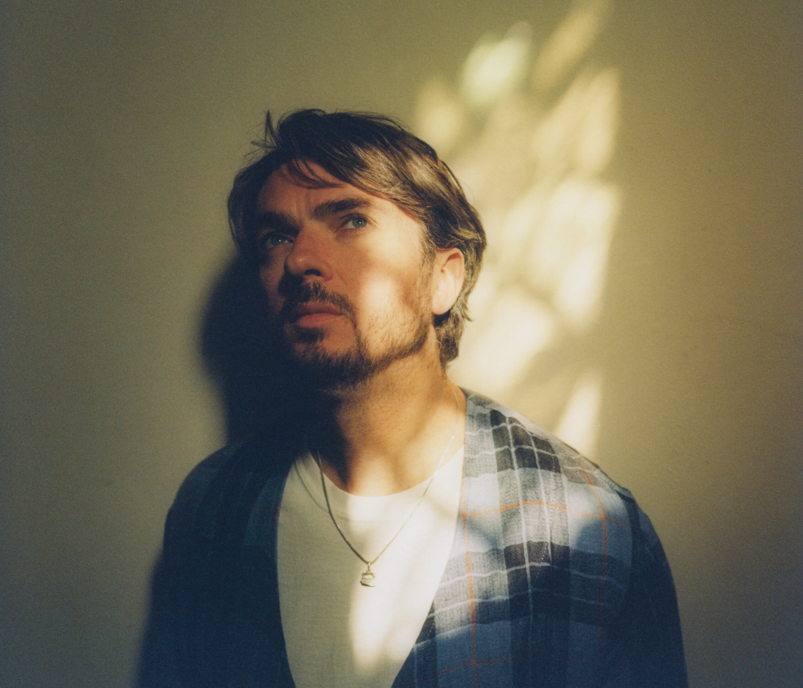 Een man met een baard en een casual shirt staat in een zachte, lichtgekleurde ruimte, terwijl schaduwpatronen op zijn gezicht vallen.