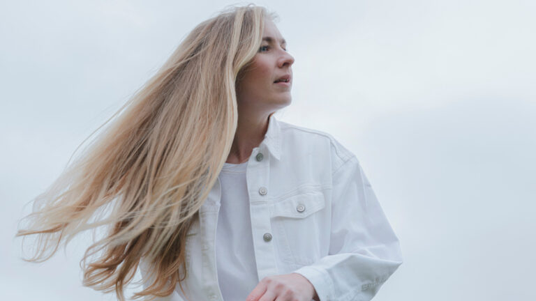 Een vrouw met lang blond haar kijkt naar de lucht, gekleed in een witte jas tegen een grijzige achtergrond.