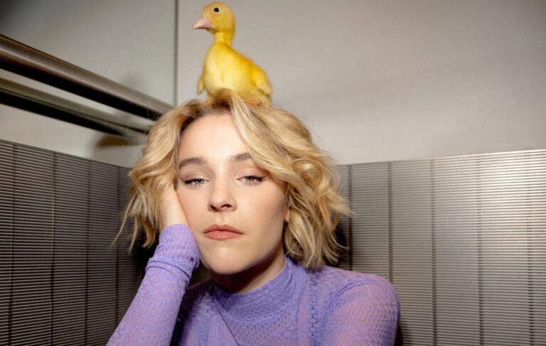 Zangeres Pommelien Thijs heeft haar hand tegen haar gezicht, ze kijkt met kleine ogen in de camera en heeft een gele eend op haar hoofd.