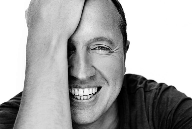 Een man lacht breed en steunt zijn hoofd op zijn arm in een zwart-witfoto.