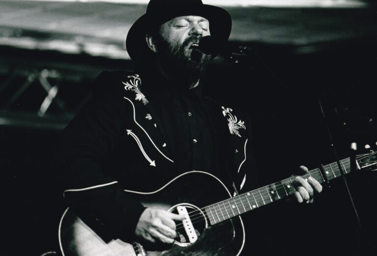 Een muzikant in zwart-wit, gekleed in een cowboyhoed en versierde jas, terwijl hij een gitaar bespeelt en zingt.