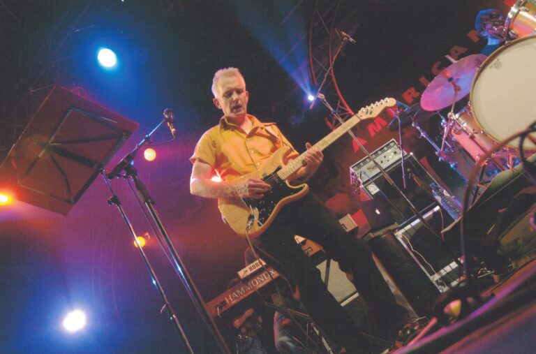 Een muzikant speelt gitaar op een live optreden met kleurrijke verlichting op de achtergrond.