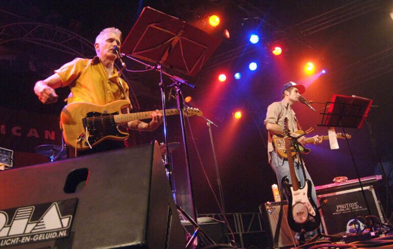 Twee muzikanten op het podium tijdens een live optreden, met kleurrijke lichten op de achtergrond.