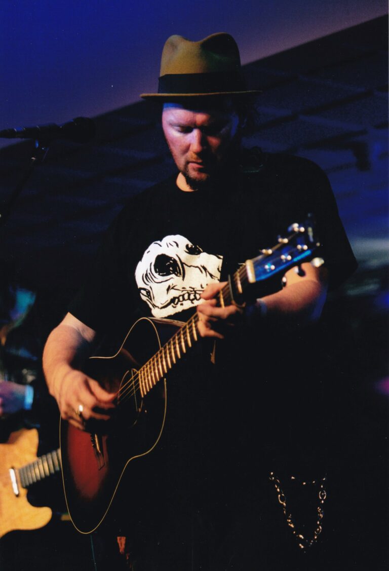 Een muzikant speelt gitaar op een podium, met een hoed en een T-shirt met een skull-print.