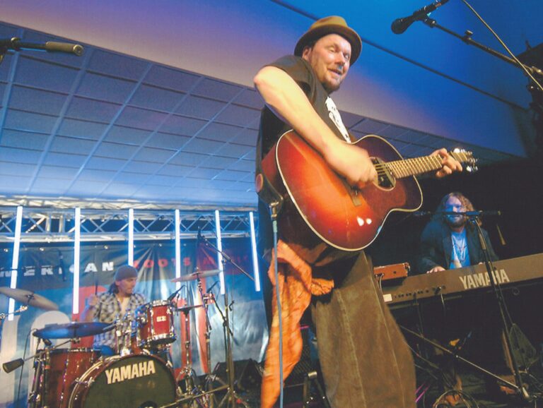 Een artiest speelt gitaar op het podium terwijl een drummer en een toetsenist op de achtergrond te zien zijn.