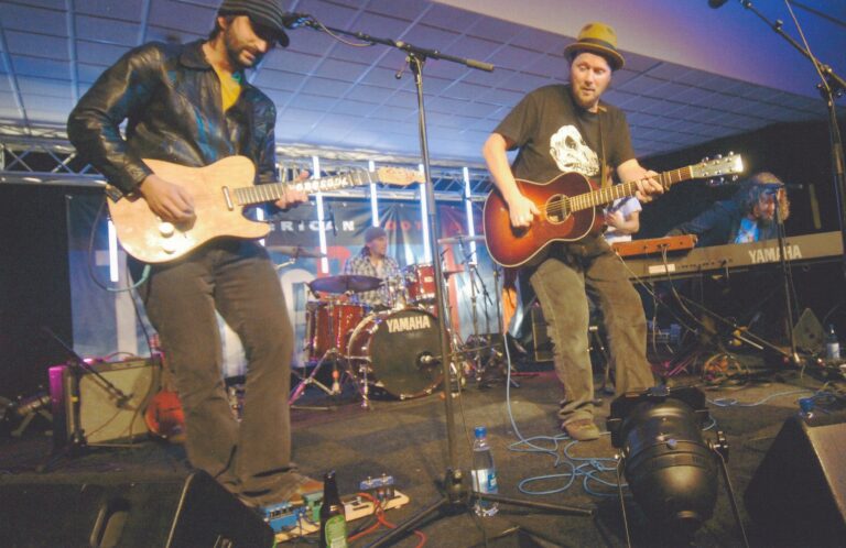Twee muzikanten spelen live op het podium, met een drummer en instrumenten op de achtergrond.