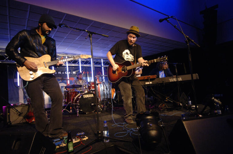 Twee muzikanten spelen live op een podium, één op een gitaar en de ander op een elektrische gitaar.