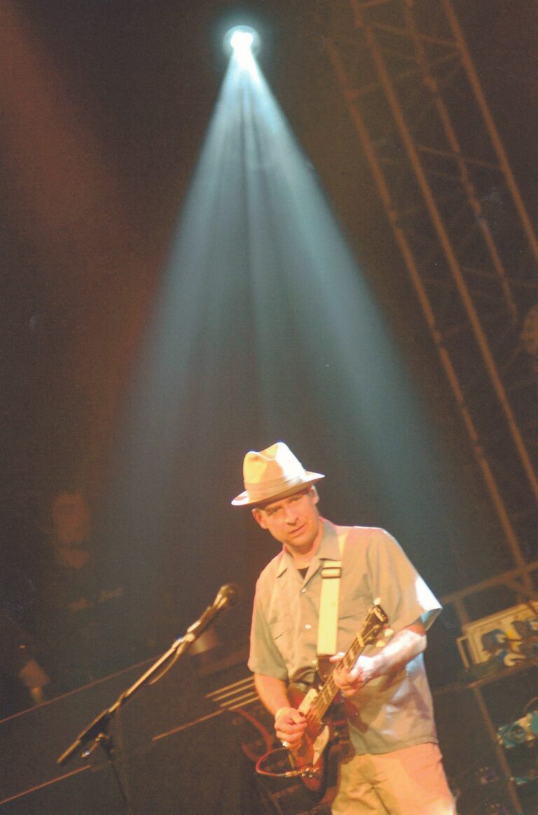 Een muzikant met een gitaar staat in een spotlight op een podium.