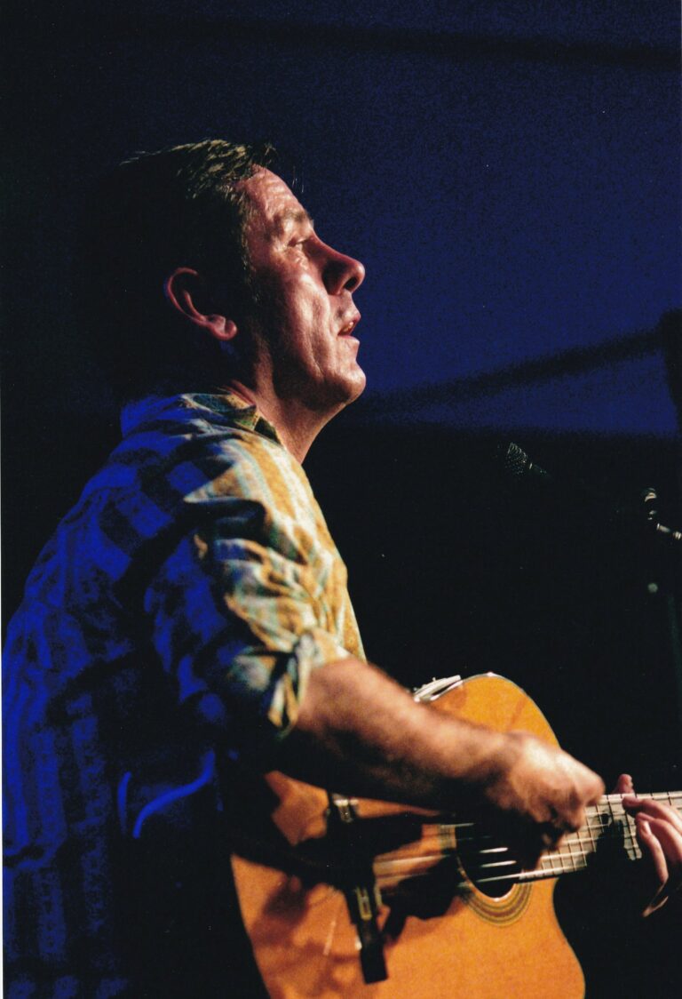 Een man speelt gitaar op een podium en kijkt naar boven in een blauwe belichting.