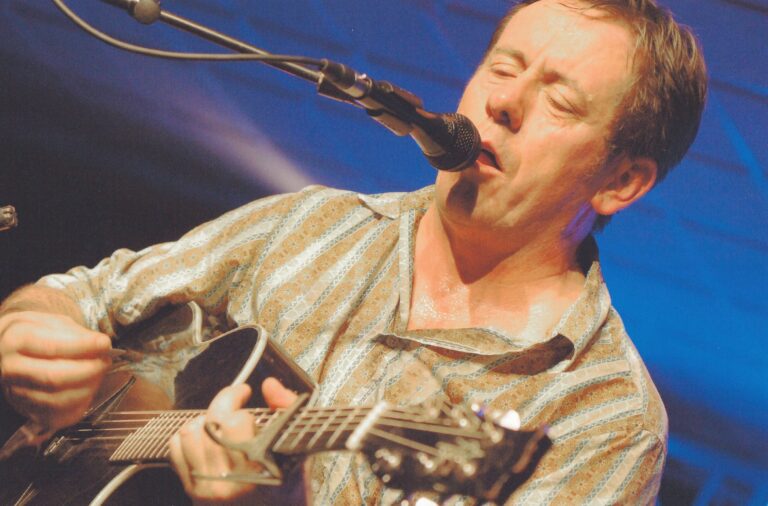 Een man speelt gitaar en zingt in een microfoon op een podium.