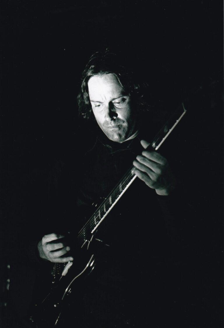 Een muzikant speelt gitaar in een donkere omgeving met opvallende verlichting op zijn gezicht.