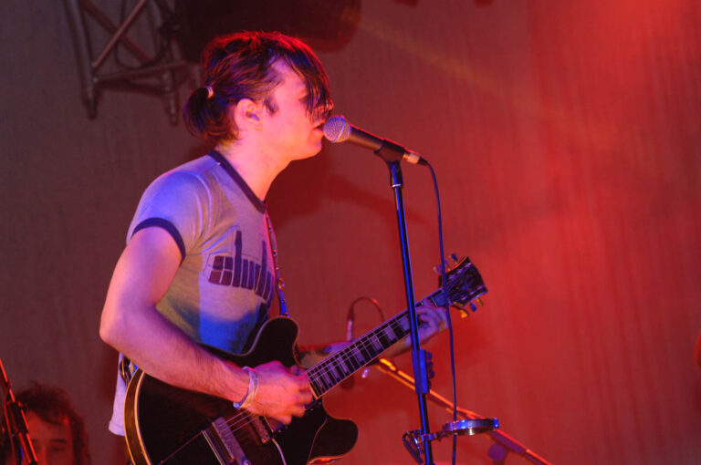 Een muzikant zingt en speelt gitaar onder kleurrijke verlichting op een podium.
