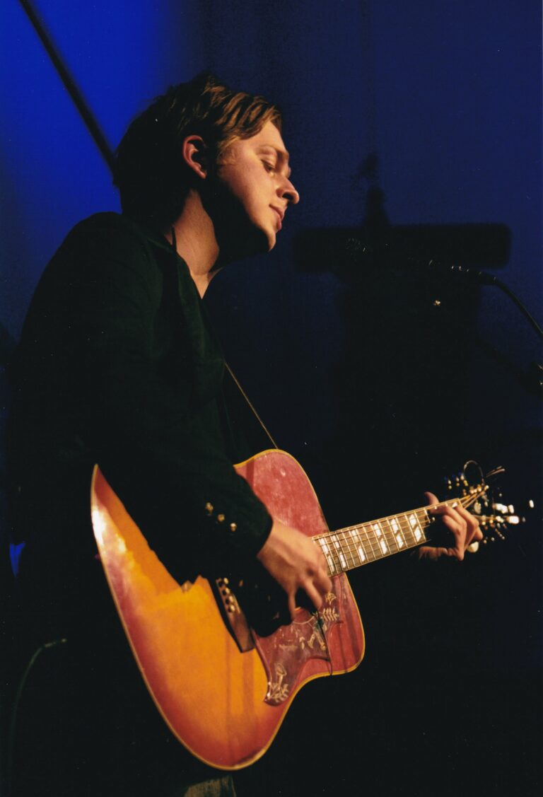 Een muzikant speelt gitaar tijdens een optreden met een lichtblauwe achtergrond.