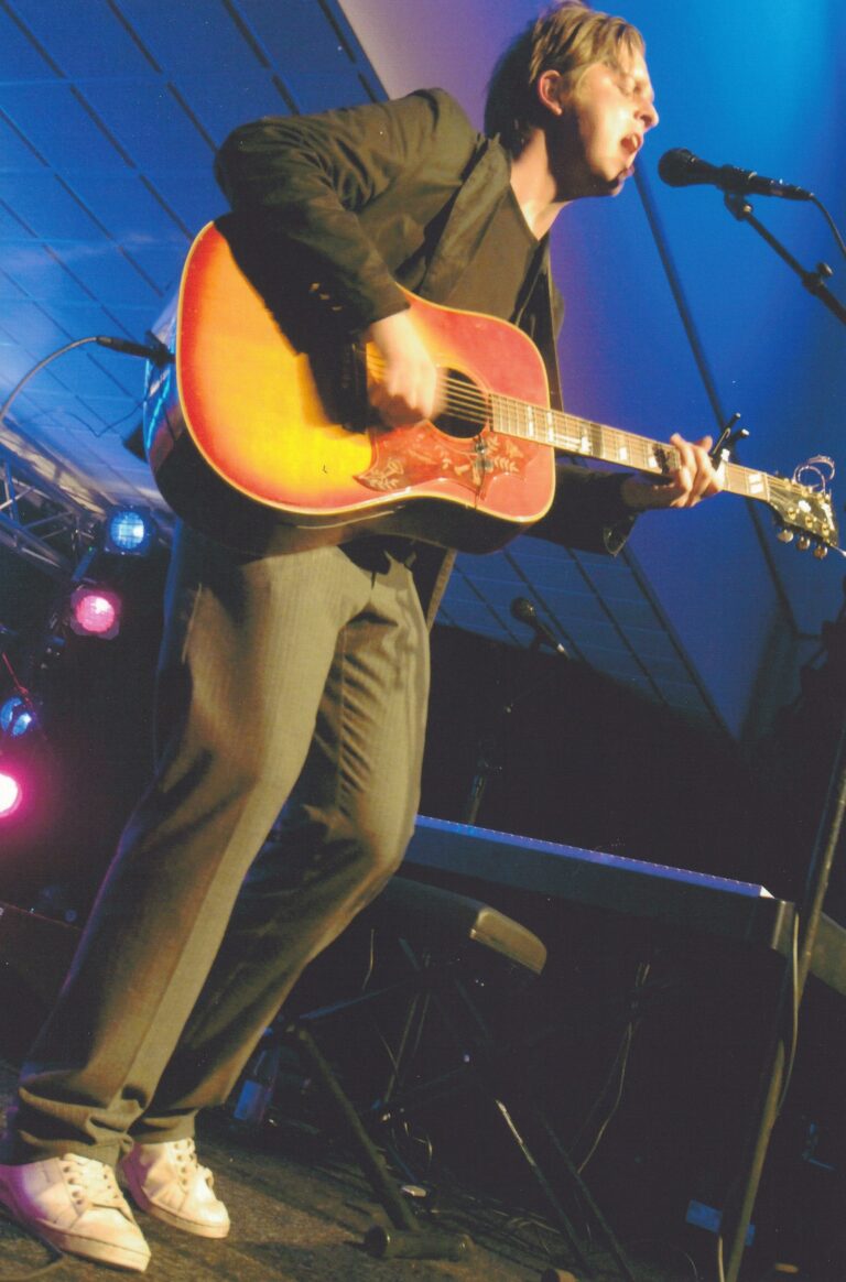 Een muzikant speelt gitaar op een podium met gekleurde lichten.