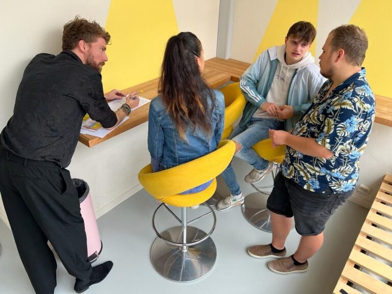 Een groep vier mensen zit aan een tafel in een lichte ruimte met gele muren, terwijl een persoon aan de andere kant van de tafel schrijft.