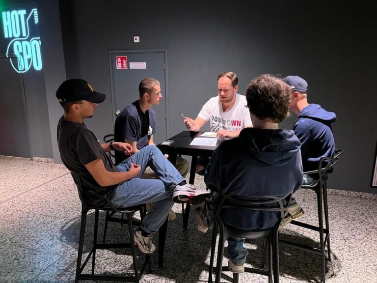 Een groep van vijf mensen zit aan een tafel en heeft een gesprek in een ruimte met een donkere achtergrond.