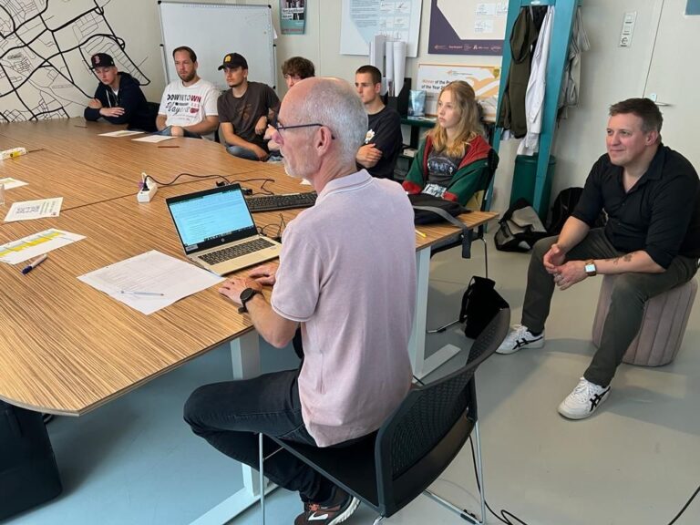 Een groep mensen is bijeen in een vergaderruimte, terwijl een man aan een laptop werkt en anderen aandachtig luisteren.