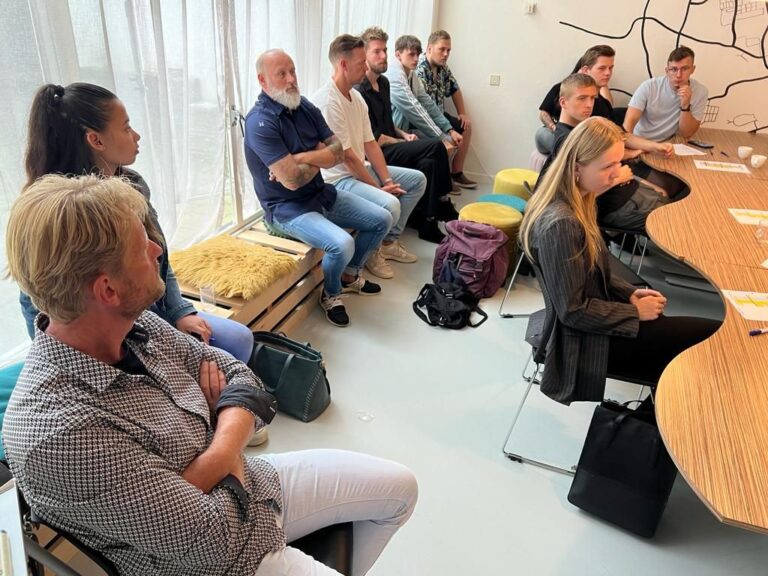 Een groep mensen zit in een vergaderruimte, met enkele personen aan een ronde tafel en anderen eromheen.