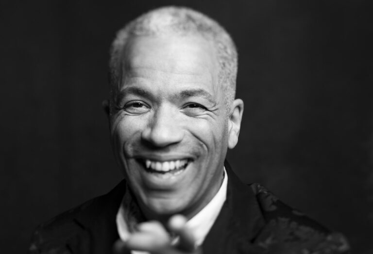 Een zwartwit portretfoto van Arnold Veeman die lachend naar de camera wijst.