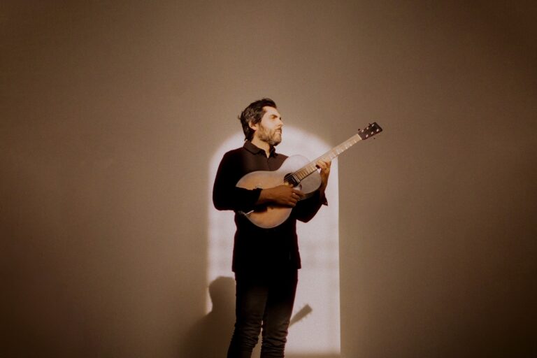 Zanger Ariel Posen staat voor een muur met een gitaar in zijn hand, hij kijkt op zij. Er valt van voren licht op hem waardoor er een schaduw op de muur ontstaat.